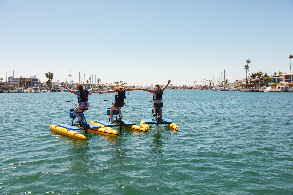 Riders pedaling hydrobikes on calm Alamitos Bay with homes and palm trees behind them