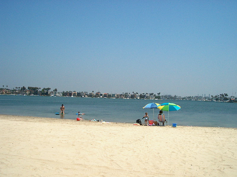 Families with beach umbrellas wading in calm, shallow water on the bay side of the Alamitos Peninsula