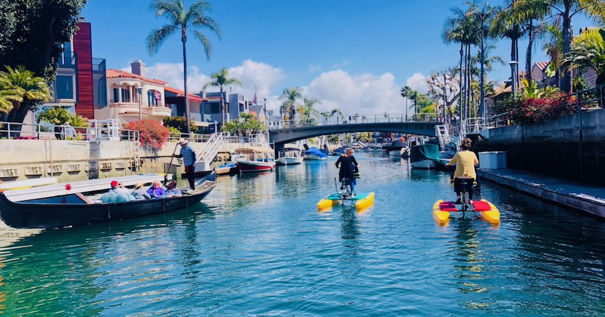 A gondolier rows guests along a Naples canal while two hydrobike riders pedal past waterfront homes and palms