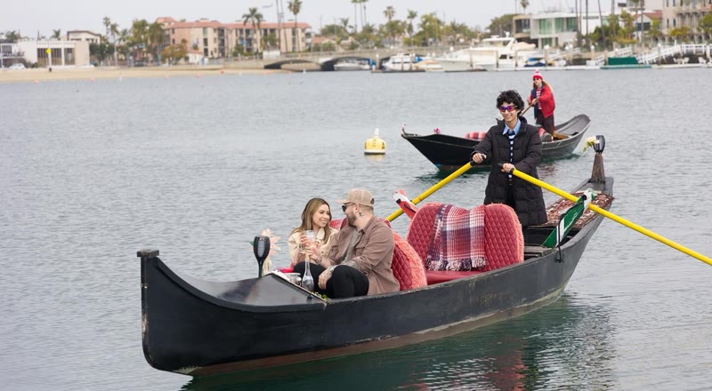 A Gondola Getaway gondolier rows a couple in a black gondola with red cushions on Alamitos Bay near Naples