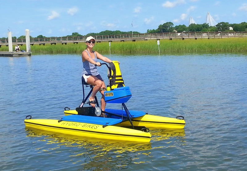 A smiling rider pedaling a yellow and blue Hydrobike on a marsh-lined waterway