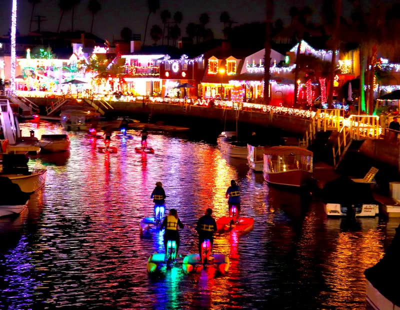 LED-lit hydrobikes in a Naples Island canal at night, surrounded by homes covered in Christmas lights.