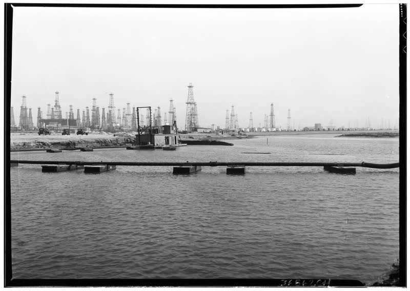 Black and white 1932 photo of a dredger and pipeline in the Marine Stadium channel, with oil derricks behind.