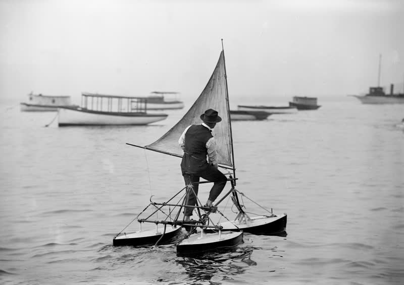 Black and white photo of a man pedaling a three-hulled water tricycle with a small sail, around 1900.