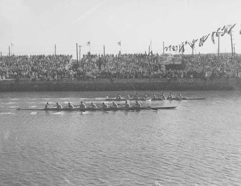 Black and white 1932 photo of eight-oared rowing crews racing past packed stands at Long Beach