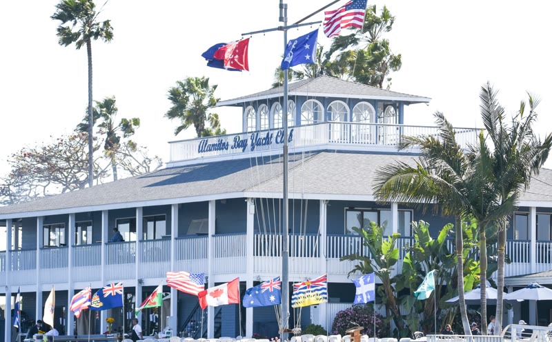 The blue-gray two-story Alamitos Bay Yacht Club clubhouse with international flags and palm trees