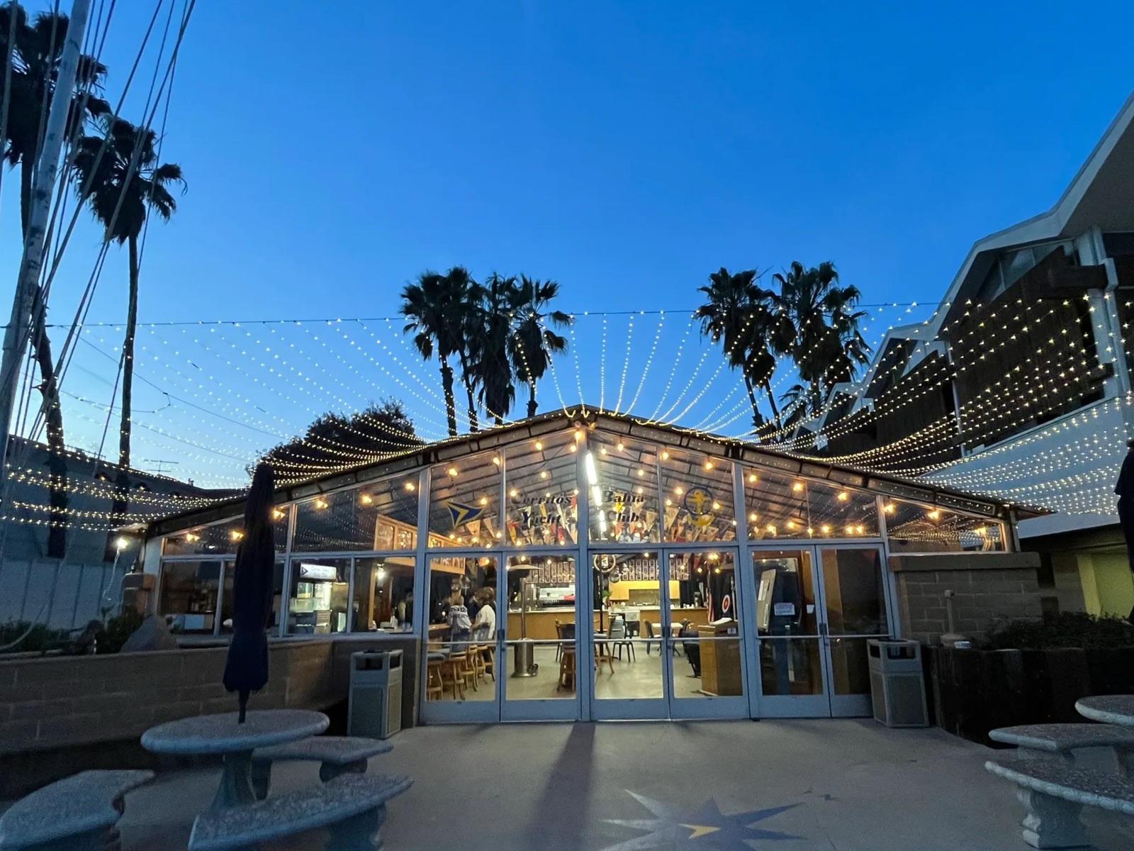 The Cerritos Bahia Yacht Club clubhouse at dusk, a glass-walled patio room glowing under string lights and palm trees