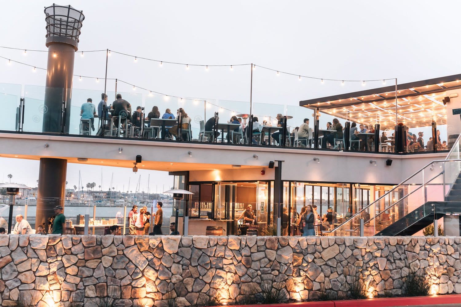 Ballast Point Brewing Long Beach at dusk, the rooftop deck full under string lights and the taproom lit up, with the marina behind
