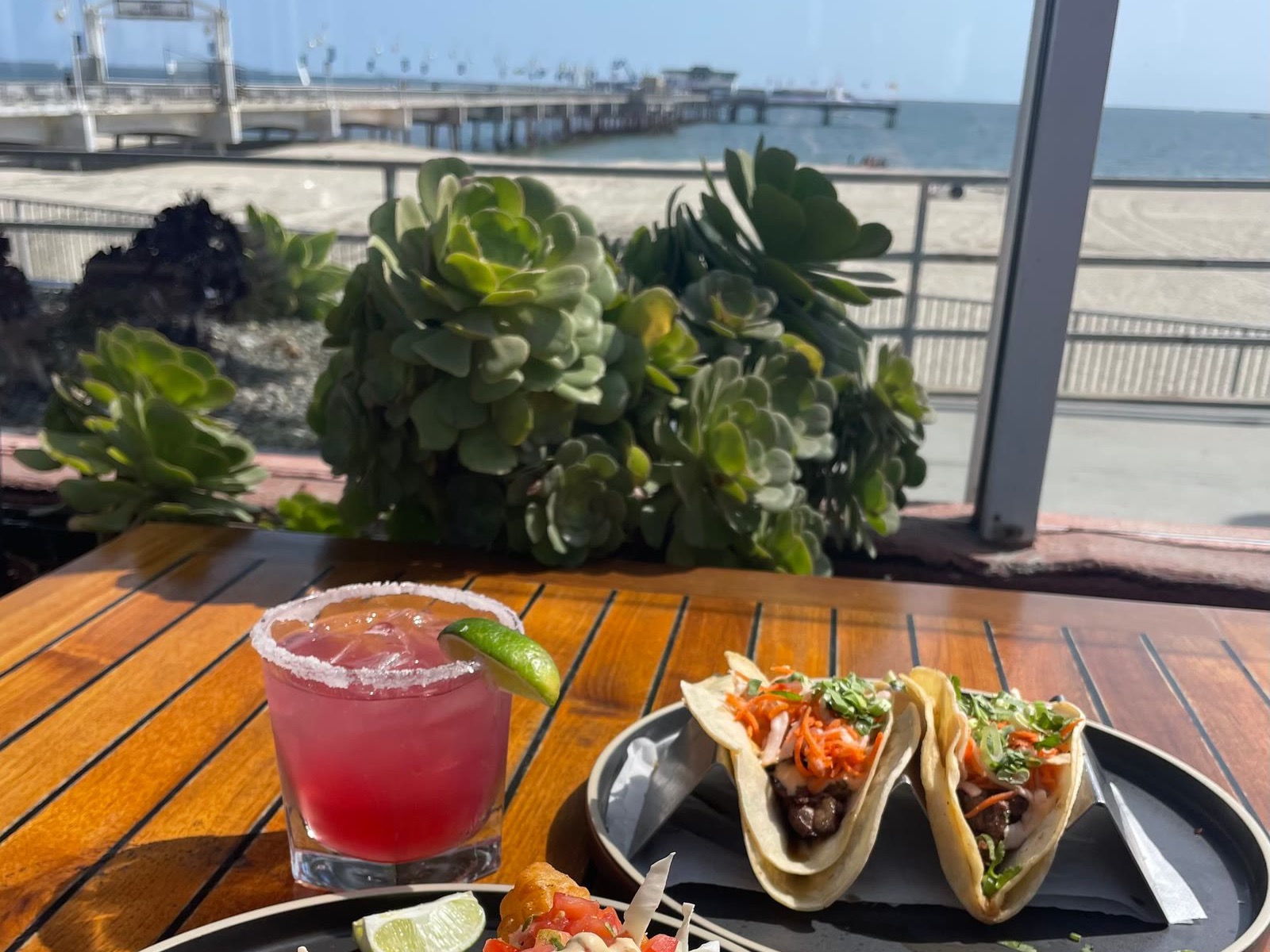 A margarita and fish tacos on the Belmont Brewing Company patio, with Belmont Pier and the beach behind on a sunny day
