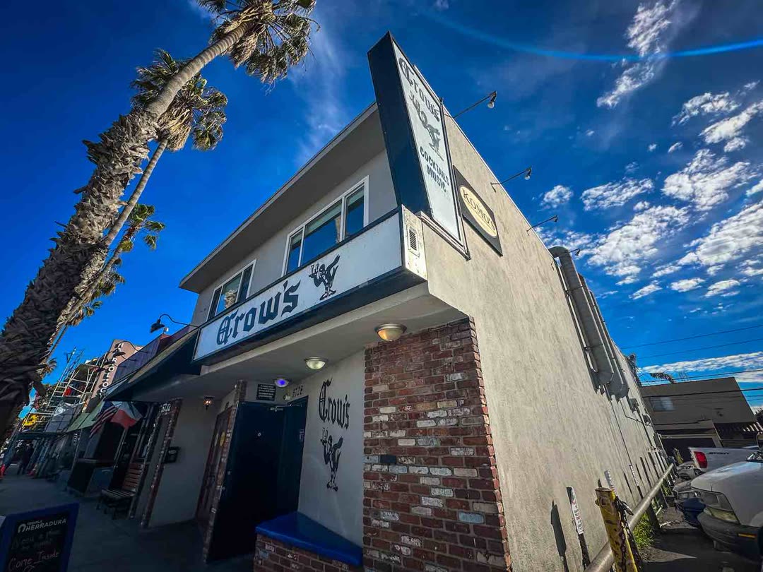 The front of Crow's Cocktails on Second Street in Naples, with its Old English style sign, brick wall and palm trees against a blue sky