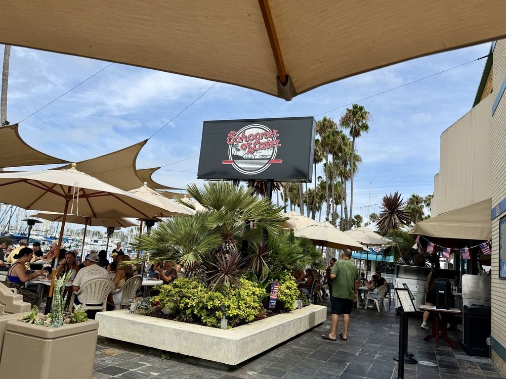 The Schooner or Later sign over the palm planter at the entrance on Marina Drive, with the umbrella-shaded patio and marina masts behind