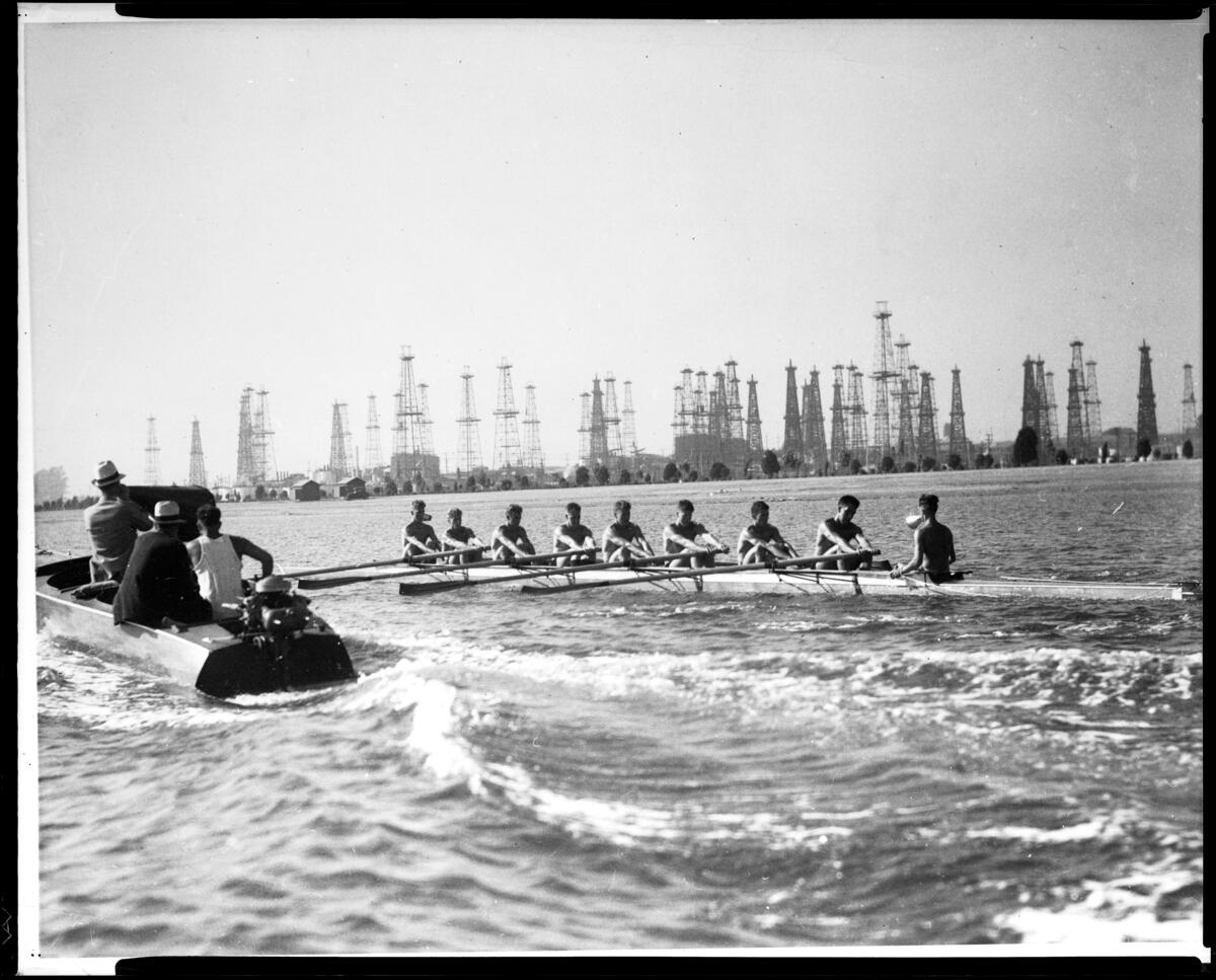 Rowing crew training on the water in Long Beach in 1932 beside a coach's launch, with oil derricks behind