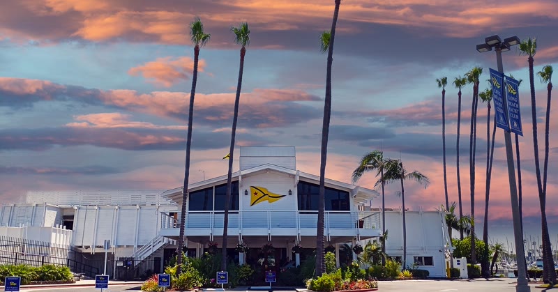 The white Long Beach Yacht Club clubhouse with its yellow burgee, tall palm trees and a colorful sky